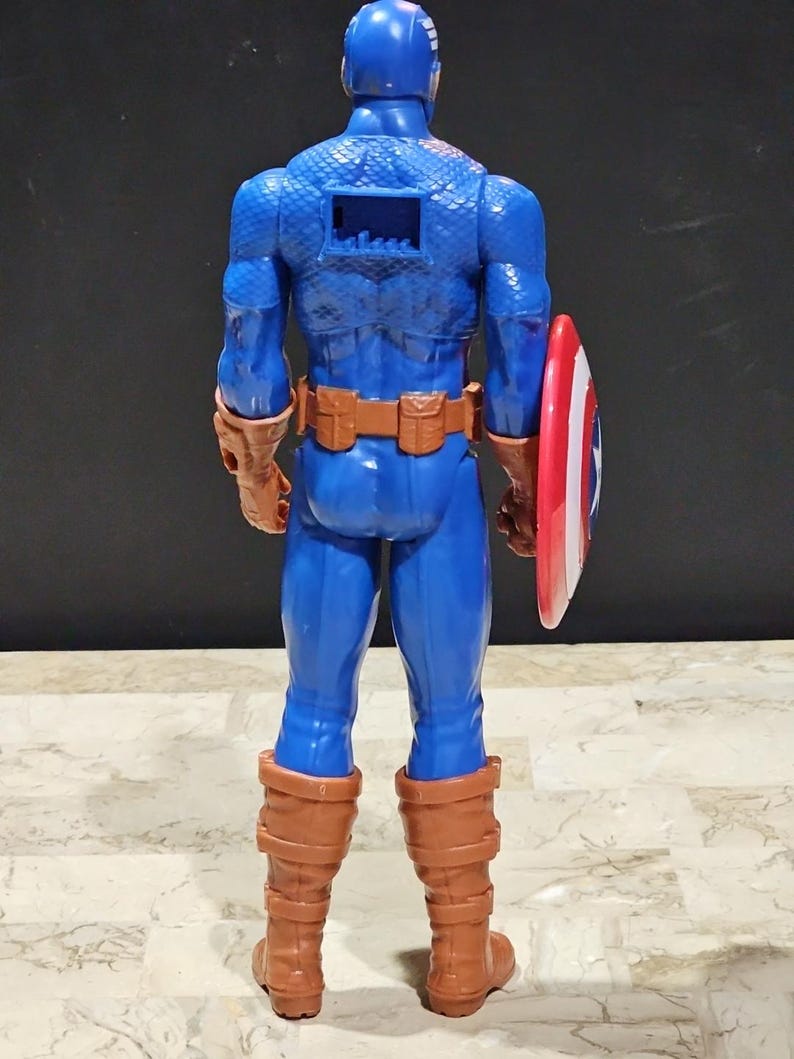 May include: A blue and red plastic action figure of Captain America, wearing a blue and red costume with a red, white, and blue shield. The figure is standing with its back to the camera, and its arms are at its sides.