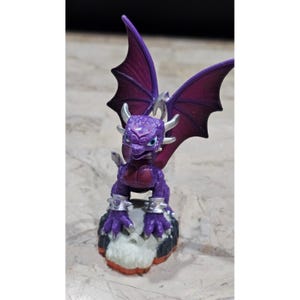 May include: A purple dragon figurine with large, dark purple wings and silver accents. The dragon has blue eyes and is perched on a rocky base with white and orange details. The figurine is a collectible toy.