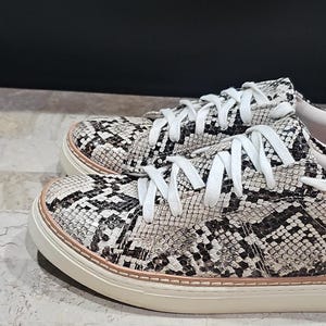 May include: A pair of low-top sneakers with a snakeskin pattern in black, white, and gray. The shoes have white laces, a white sole, and a light pink trim around the sole.