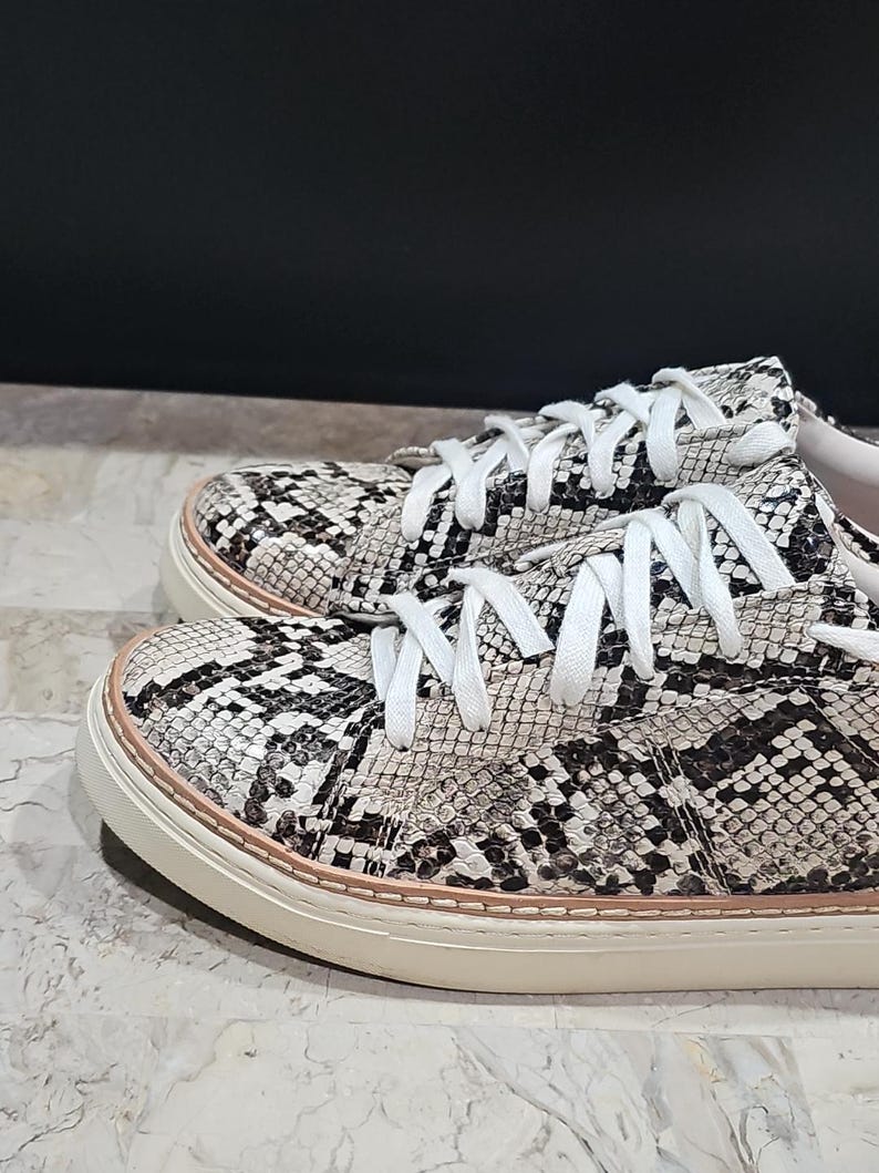 May include: A pair of low-top sneakers with a snakeskin pattern in black, white, and gray. The shoes have white laces, a white sole, and a light pink trim around the sole.