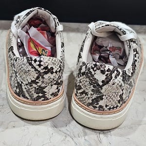 May include: A pair of snake-print sneakers. The shoes feature a white, black, and tan pattern with a light pink trim around the sole. The soles are white, and the interior is white with paper stuffing.