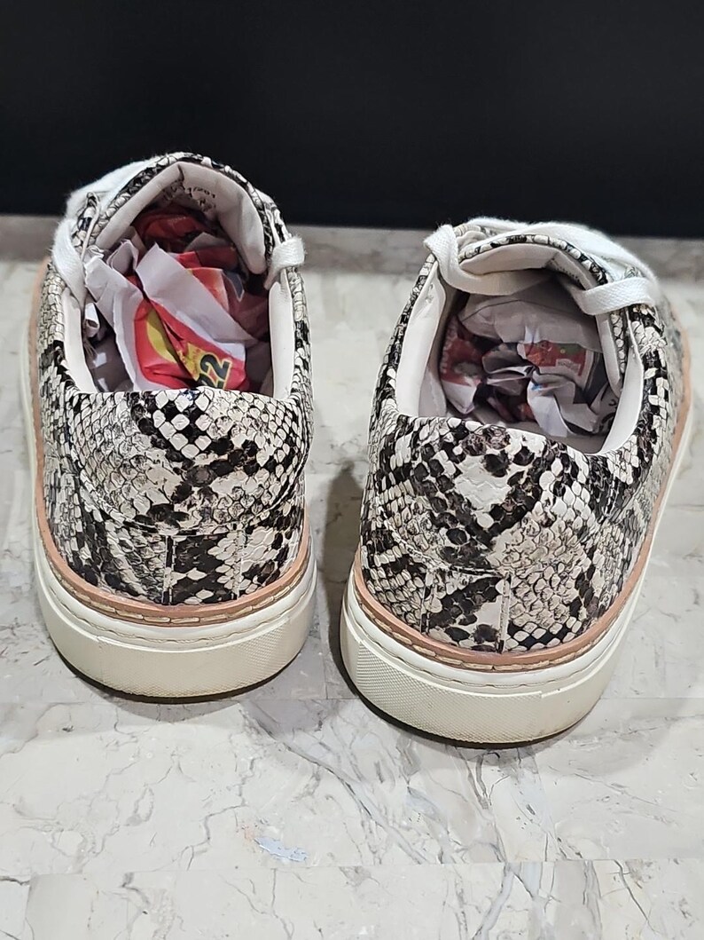 May include: A pair of snake-print sneakers. The shoes feature a white, black, and tan pattern with a light pink trim around the sole. The soles are white, and the interior is white with paper stuffing.