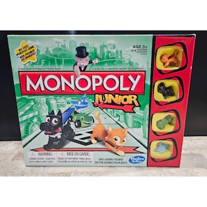 May include: Monopoly Junior board game box. The box is green, red, and white, with the Monopoly Junior logo and illustrations of game pieces. The game is for 2-4 players, ages 5+. Includes game pieces such as a dog, cat, car, and other tokens.