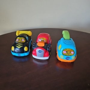 May include: Three toy cars with light-up faces. The cars are black, red, and blue with yellow, orange, and green accents. The cars are VTech brand.
