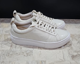 May include: A pair of off-white, glossy sneakers with white laces and soles. The shoes have a low-top design and a smooth, shiny finish. The sneakers are displayed on a marble-patterned surface against a dark background.