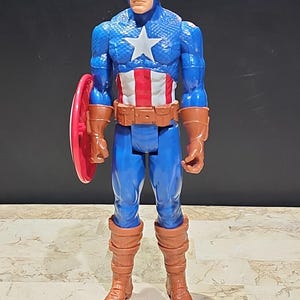 May include: A plastic action figure of Captain America, wearing his iconic red, white, and blue costume. He is holding his red, white, and blue shield in his left hand. The figure is approximately 12 inches tall.