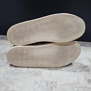 May include: A pair of sneakers with white soles and a textured, patterned upper. The soles are off-white with a worn appearance. The shoes are set against a dark background and a light-colored surface.