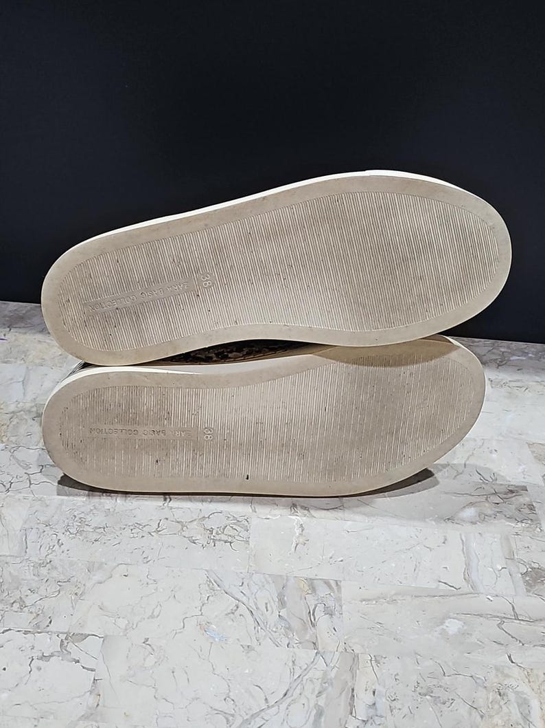 May include: A pair of sneakers with white soles and a textured, patterned upper. The soles are off-white with a worn appearance. The shoes are set against a dark background and a light-colored surface.