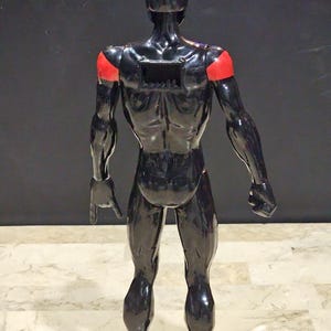 May include: A black, glossy figurine of a human form, with red accents on the shoulders. The figure is posed with its back to the viewer, showcasing muscular definition. The figurine has a rectangular opening on its back.