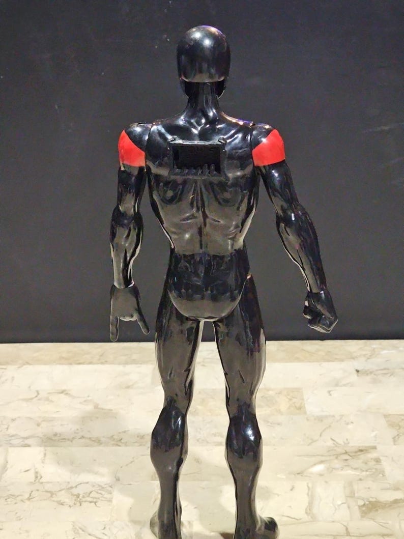 May include: A black, glossy figurine of a human form, with red accents on the shoulders. The figure is posed with its back to the viewer, showcasing muscular definition. The figurine has a rectangular opening on its back.