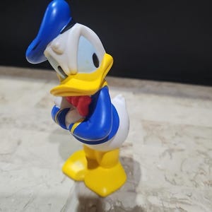 May include: A yellow and blue plastic figurine of Donald Duck wearing a sailor hat. The figurine is standing with its arms crossed and is looking to the side.