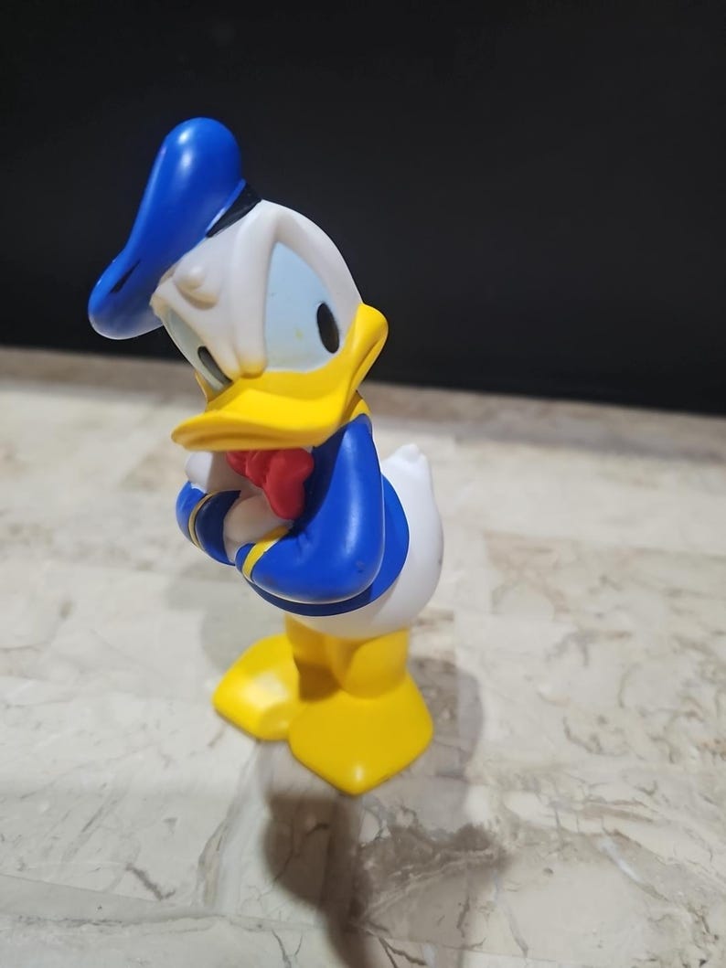 May include: A yellow and blue plastic figurine of Donald Duck wearing a sailor hat. The figurine is standing with its arms crossed and is looking to the side.