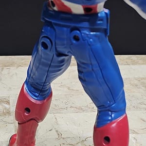 May include: A blue and red action figure of Captain America with articulated legs and arms. The figure is posed with its legs slightly apart and its arms bent at the elbows.