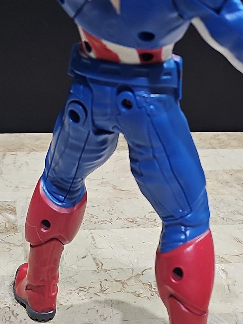 May include: A blue and red action figure of Captain America with articulated legs and arms. The figure is posed with its legs slightly apart and its arms bent at the elbows.