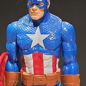 May include: A blue and red action figure of Captain America. The figure has a blue and white star on the chest, a red, white, and blue belt, and a brown leather-like arm band. The figure is wearing a blue helmet with a white "A" and a white wing on the side.