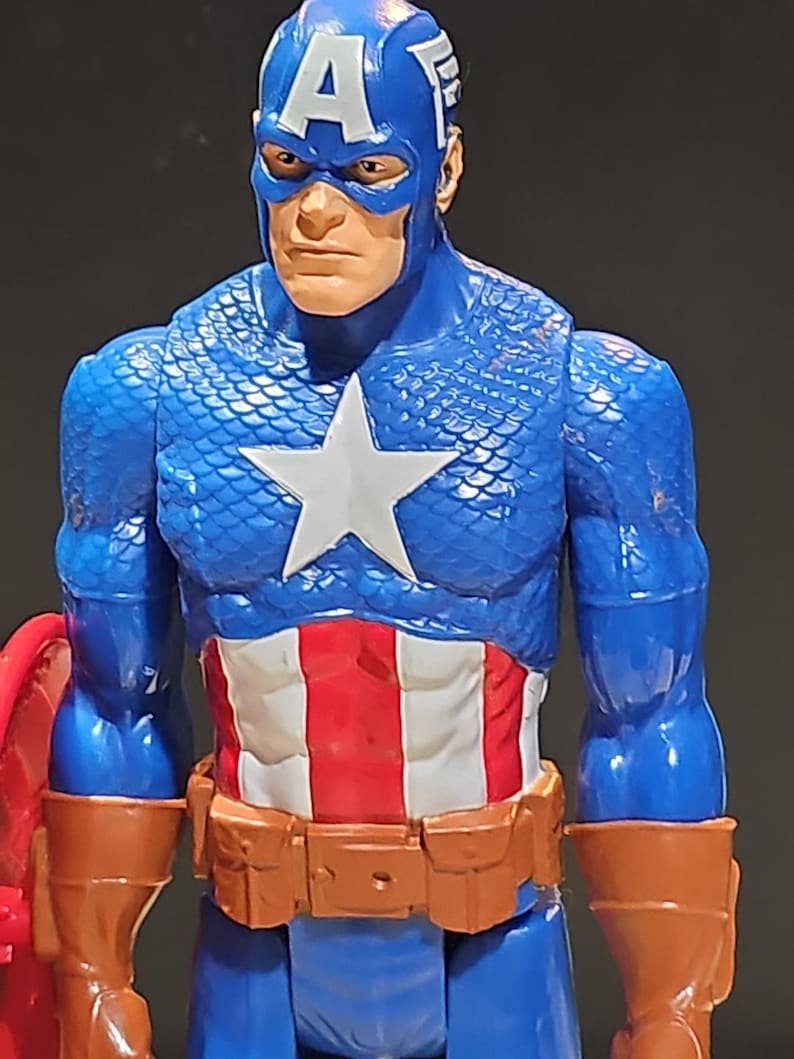 May include: A blue and red action figure of Captain America. The figure has a blue and white star on the chest, a red, white, and blue belt, and a brown leather-like arm band. The figure is wearing a blue helmet with a white "A" and a white wing on the side.