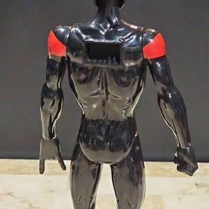 May include: A black action figure with red shoulder accents. The figure is glossy and muscular, with a rectangular opening on its back. The figure is posed with one hand open and the other clenched into a fist.