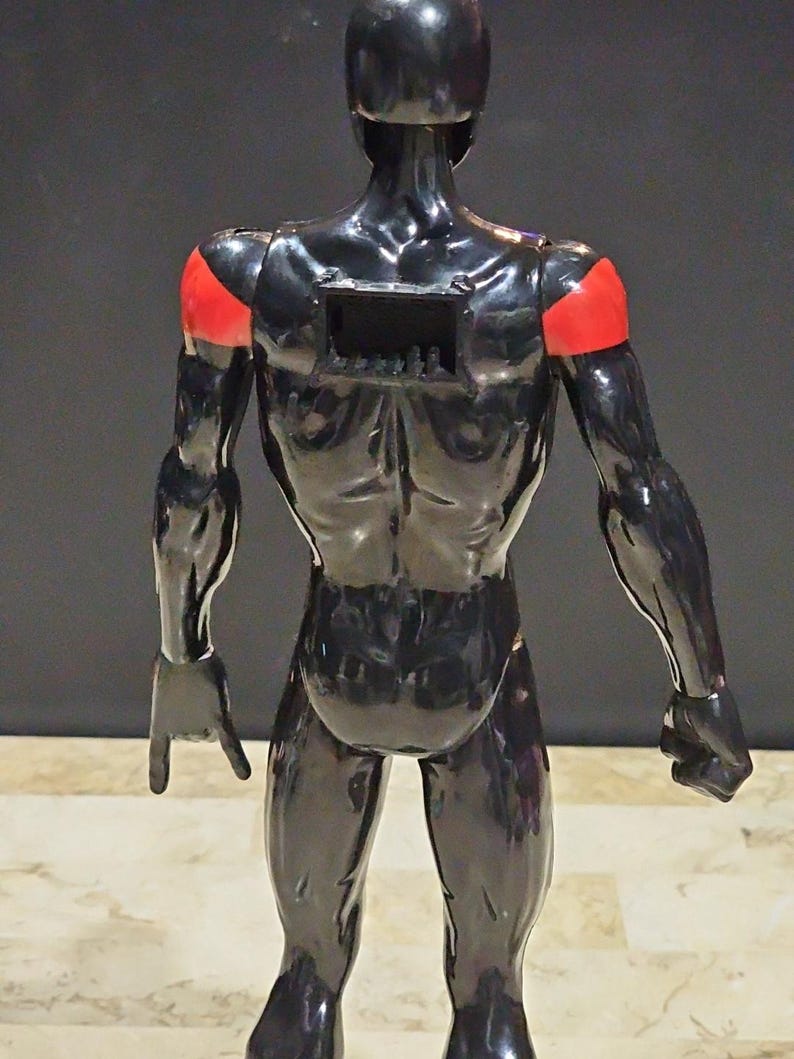 May include: A black action figure with red shoulder accents. The figure is glossy and muscular, with a rectangular opening on its back. The figure is posed with one hand open and the other clenched into a fist.