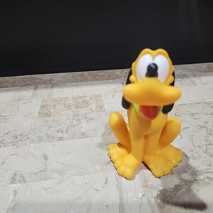 May include: A yellow plastic toy figurine of Pluto, a cartoon dog from Disney. Pluto is sitting with his tongue sticking out and has black ears and a green collar.