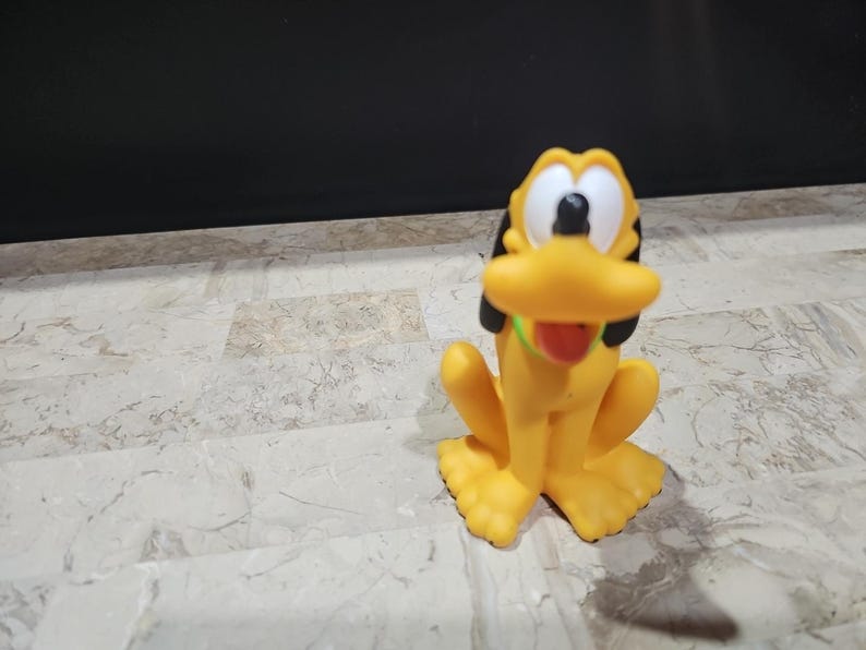 May include: A yellow plastic toy figurine of Pluto, a cartoon dog from Disney. Pluto is sitting with his tongue sticking out and has black ears and a green collar.