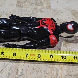 May include: A black and red Spider-Man action figure, approximately 11 inches long, lies on a surface. The figure has a red spider emblem on the chest and red accents on the arms. The head is black with white eye details.