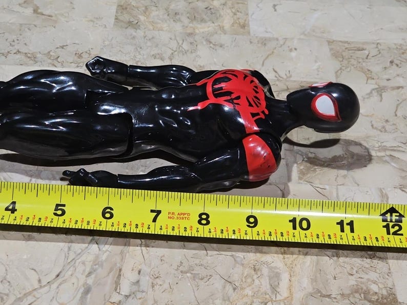 May include: A black and red Spider-Man action figure, approximately 11 inches long, lies on a surface. The figure has a red spider emblem on the chest and red accents on the arms. The head is black with white eye details.