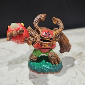 May include: A brown and red tree-like figure with a green base. The figure has a red hand with a white claw and a brown arm. The figure has a brown head with a red face and a black mask.