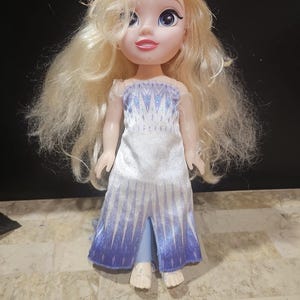 May include: A small doll with long blonde hair wearing a white and blue dress with a snowflake pattern. The doll has blue eyes and a pink mouth.
