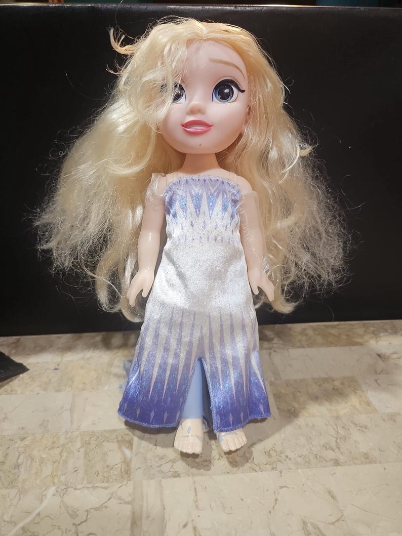 May include: A small doll with long blonde hair wearing a white and blue dress with a snowflake pattern. The doll has blue eyes and a pink mouth.