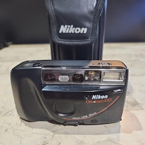 Pode incluir: Câmera Nikon One-Touch 100 preta de 35 mm com uma capa preta combinando. A câmera tem uma lente e flash prateados, com o logotipo Nikon e o nome do modelo em branco e vermelho. A capa tem o logotipo Nikon.