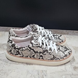 May include: A pair of sneakers with a snakeskin pattern in black, white, and gray. The shoes have white laces and a cream-colored sole. The sneakers are casual and stylish.