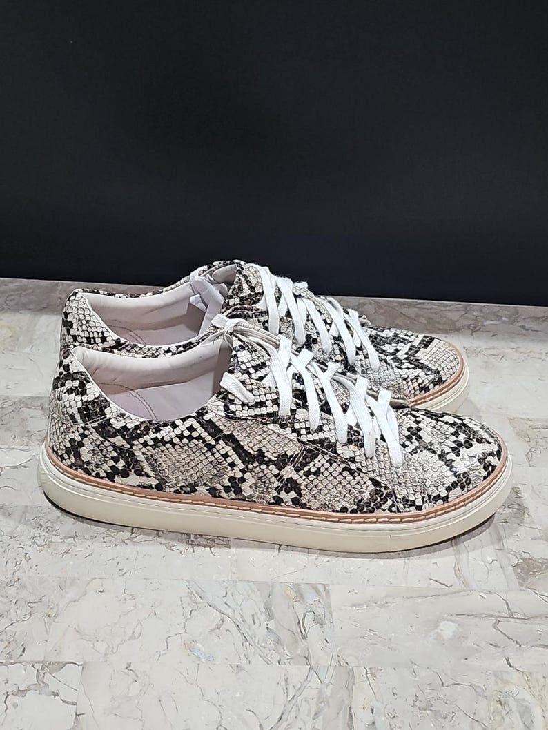May include: A pair of sneakers with a snakeskin pattern in black, white, and gray. The shoes have white laces and a cream-colored sole. The sneakers are casual and stylish.