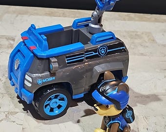 May include: A blue and gray toy vehicle with a rotating light on top. The vehicle has a paw print logo on the side. A brown and blue toy dog figure stands next to the vehicle.