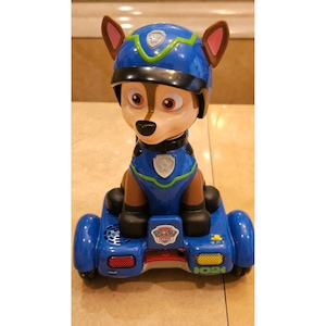 May include: A blue and brown Chase toy from the Paw Patrol series. The toy features a blue helmet and uniform, with the Paw Patrol logo on the front. The toy is mounted on a blue vehicle with the number 024.