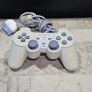 Op de afbeelding: Een witte Sony PlayStation 1-controller met een grijze kabel. De controller heeft een grijze analoge stick, een zwart D-pad en zwarte knoppen met de labels 'Start', 'Select', 'Driehoek', 'Cirkel', 'Kruis' en 'Vierkant'.