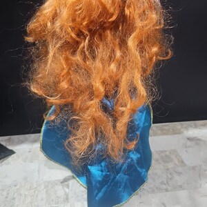 Puede incluir: Una mu&ntilde;eca con el pelo rojo y rizado que lleva una capa azul con ribetes dorados. La mu&ntilde;eca est&aacute; de espaldas a la c&aacute;mara.