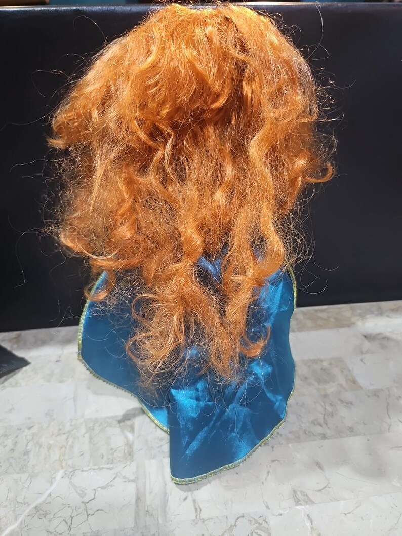 Puede incluir: Una mu&ntilde;eca con el pelo rojo y rizado que lleva una capa azul con ribetes dorados. La mu&ntilde;eca est&aacute; de espaldas a la c&aacute;mara.