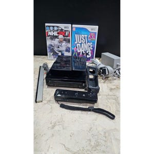 Puede incluir: Una consola Wii negra con dos carcasas de juegos, un mando a distancia y otros accesorios. Las carcasas de los juegos son para "NHL 2K11" y "Just Dance 3". La consola está sobre una superficie clara.