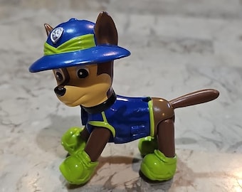 May include: A plastic toy figure of a cartoon dog, wearing a blue hat and uniform with green accents. The dog has brown fur and is posed on all fours, with a tail extending from its rear.