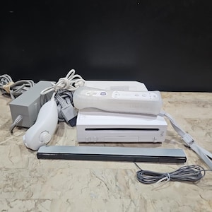 May include: A white Nintendo Wii console with a white Wii remote, a white nunchuck, a black Wii sensor bar, and a white power adapter. The console is on a light brown surface.
