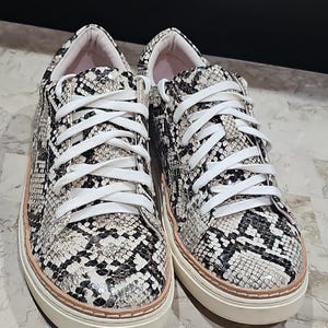 May include: A pair of snake-print sneakers with white laces. The shoes have a cream-colored sole with a light pink trim. The uppers feature a pattern of black, gray, and cream, resembling snakeskin. The sneakers are new and unworn.