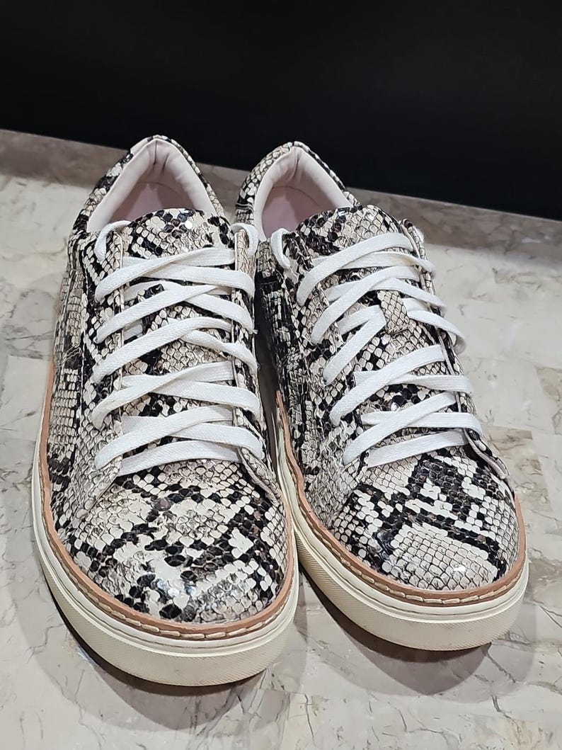 May include: A pair of snake-print sneakers with white laces. The shoes have a cream-colored sole with a light pink trim. The uppers feature a pattern of black, gray, and cream, resembling snakeskin. The sneakers are new and unworn.