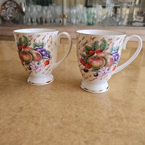 May include: Two white ceramic mugs with gold trim and a floral design featuring apples, grapes, and berries. The mugs have handwritten text on the side.