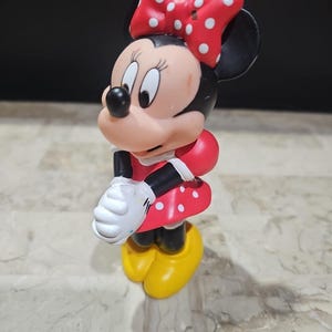 May include: A red and white polka dot dress with a red bow on top of the head. The figure is a plastic figurine of Minnie Mouse.