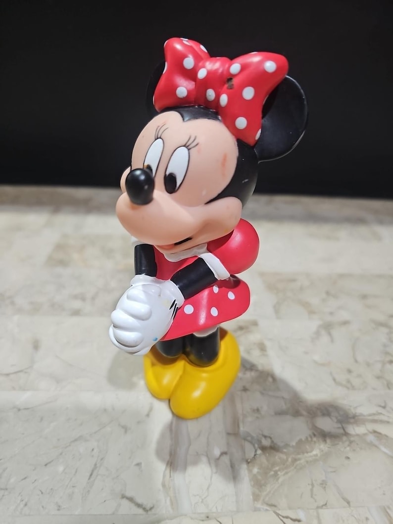 May include: A red and white polka dot dress with a red bow on top of the head. The figure is a plastic figurine of Minnie Mouse.