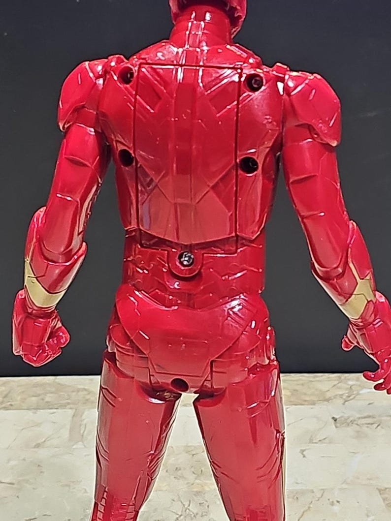 Pu&ograve; includere: Figurina di azione in plastica rossa di Iron Man, in piedi con le braccia distese, vista da dietro. La figura ha una tuta rossa dettagliata, testurizzata, con accenti dorati.