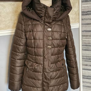 Könnte beinhalten: Eine braune gesteppte Jacke mit Kapuze und hohem Kragen. Die Jacke hat einen Reißverschluss und Knopf-Akzente.