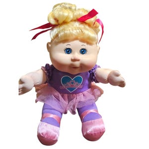 May include: A Cabbage Patch Kids doll dressed as a ballerina. The doll has blonde hair styled in a bun with red ribbons. It wears a purple leotard with a heart and crown design, a pink tutu, and pink ballet slippers. The doll has blue eyes.