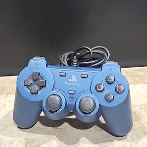 Könnte beinhalten: Ein blauer PlayStation 2-Controller mit einem schwarzen Kabel. Der Controller hat ein PlayStation-Logo und den Schriftzug "ANALOG" aufgedruckt.