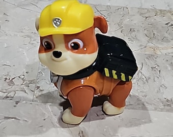 May include: A toy figure of a cartoon dog in a construction outfit. The dog has brown and white fur, wearing a yellow hard hat and a black backpack. The figure is standing on a light-colored surface.